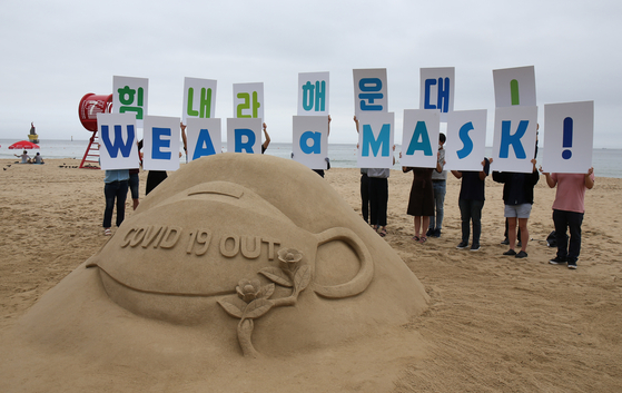Campaigners hold cards reading "Wear a mask" on Haeundae Beach, one of the most visited beaches in Busan, on July 22, behind a sand sculpture with the same message. From July 25, visitors to five main beaches in Busan including Haeundae are required to wear a mask and will be fined a maximum of 3 million won ($2,500) if spotted without one. [YONHAP]