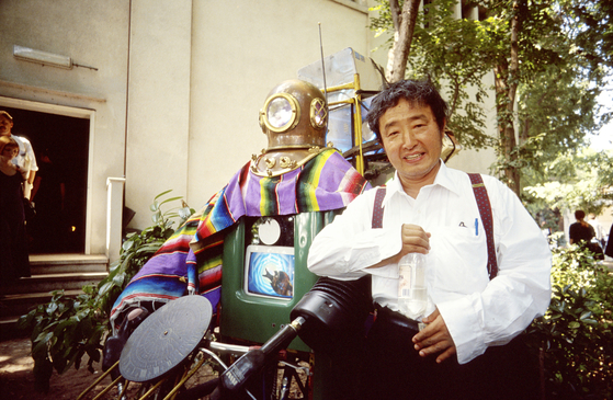 Nam June Paik poses in front of "Rehabilitation of Genghis Khan" (1993) outdoors, wearing his signature outfit: a white shirt and black trousers with suspenders. [AGE FOTOSTOCK]