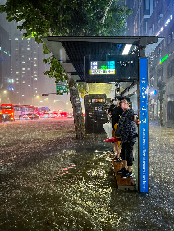 People stand on the bench of a bus station to avoid being submerged in rainwater while waiting for a bus in Seocho District, southern Seoul, Monday. [MICHAEL LEE]