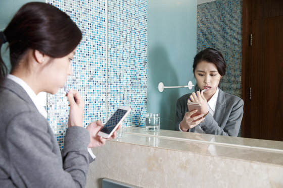 A woman scrolls her phone while brushing her teeth at an office restroom. [GETTY IMAGES]