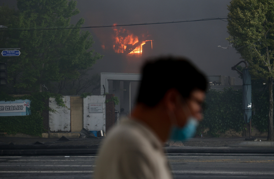 A passerby wearing a mask walks past Kumho Tire’s Gwangju plant in Songjeong-dong, Gwangsan District, as smoke rises from the fire on May 17. [YONHAP]