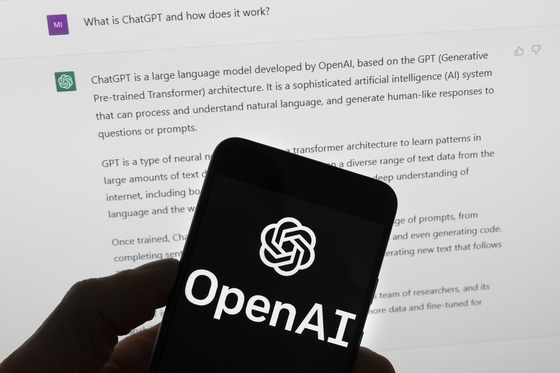 The OpenAI logo is seen on a mobile phone in front of a computer screen which displays output from ChatGPT on March 21, 2023. [AP/YONHAP]