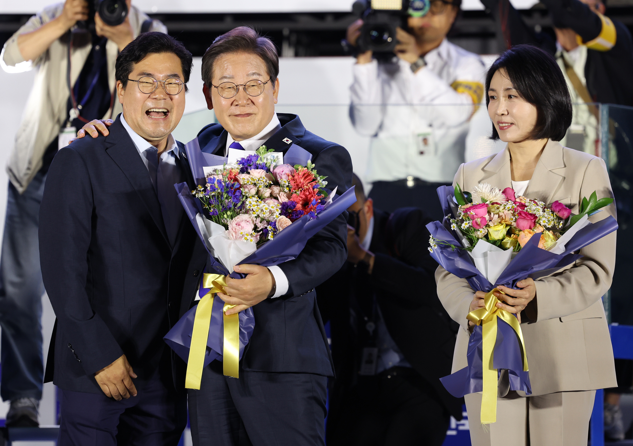 Lee Jae Myung, center, presidential candidate of the Democratic Party and presumed winner of Korea’s 21st presidential election, and his wife Kim Hae Kyung, right, receive a bouquet from Park Chan-dae, chief campaign coordinator, during a public vote-count broadcast event hosted by the party near the National Assembly in Yeouido, Seoul, on the morning of June 4. [JOINT PRESS CORPS]