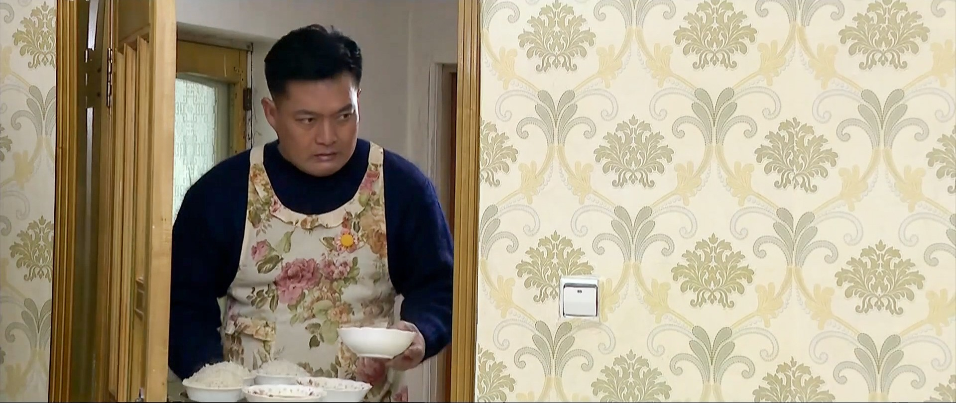 A man in the North Korean drama “A New Spring in the Field of Manchurian Cranes” wears a floral apron as he serves a meal to his wife and daughter. [KOREAN CENTRAL TELEVISION] 
