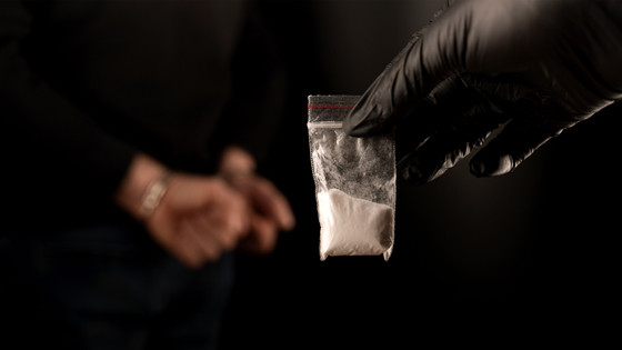 A gloved hand holds a small plastic bag of white powder as a handcuffed individual stands in the background. [JOONGANG ILBO]