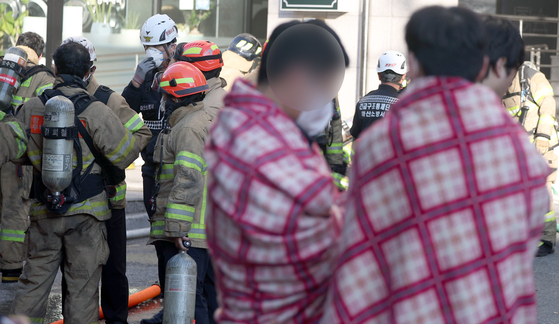 Residents of an officetel where a fire broke out from a charging EV wait in blankets provided by fire authorities as firefighters extinguish the fire at the officetel in Changwon, South Gyeongsang on Oct. 29. [YONHAP]