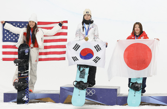 Choi Ga-on, center, who scored 90.25 points to win gold in the women’s snowboard halfpipe final at the Milano-Cortina 2026 Winter Olympics at Livigno Snow Park in Italy, stands on the podium holding up the national flag on Feb. 12. Chloe Kim, left, won silver and Mitsuki Ono, right, took bronze. [YONHAP]