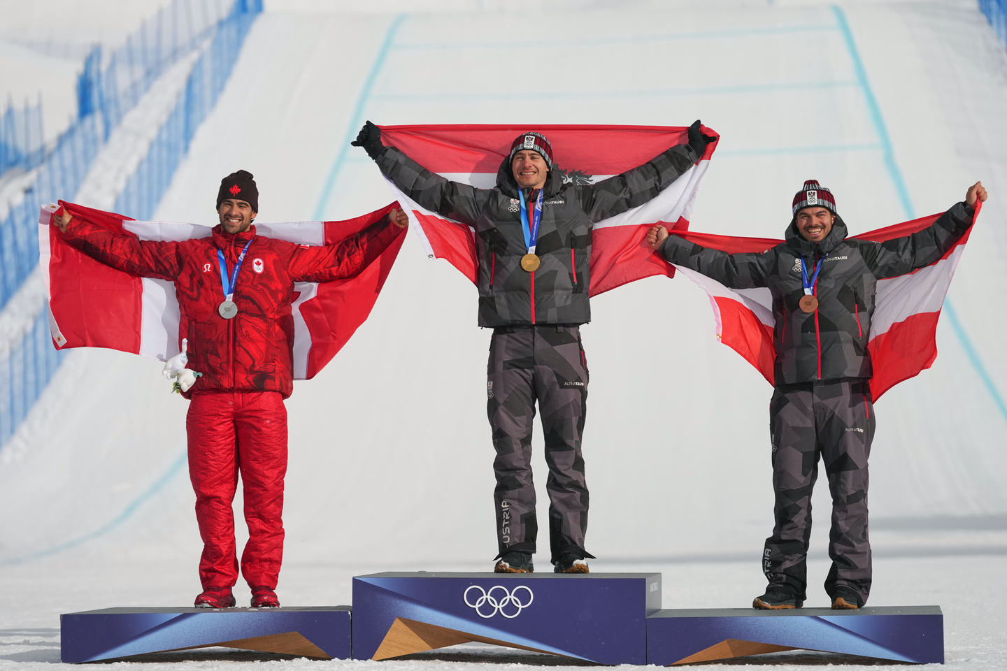 Silver medalist Canada's Eliot Grondin, left, gold medalist Austria's Alessandro Haemmerle, center, and bronze medalist Austria's Jakob Dusek celebrate with flags during the men's snowboard cross finals at the 2026 Winter Olympics, in Livigno, Italy, on Feb. 12.  [AP/YONHAP]