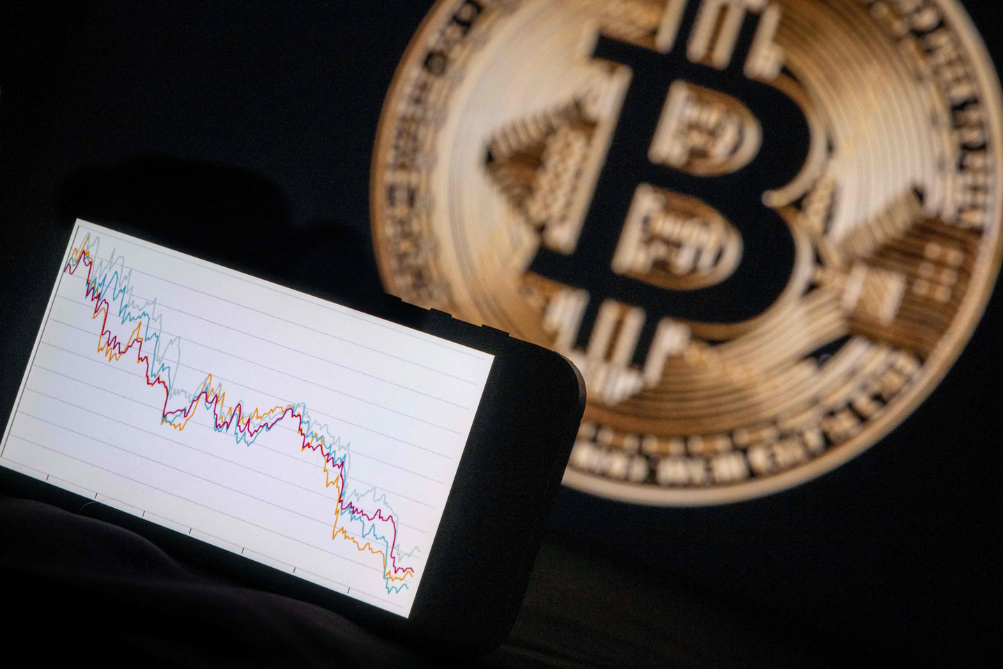 This photograph, taken in Mulhouse on Dec. 5, 2025, shows a phone displaying a downward trend in the stock market in front of a screen displaying a Bitcoin logo. The hype surrounding cryptocurrencies, fueled by the return of U.S. President Donald Trump and the hope of a pro-crypto shift in Washington, seems to have stalled with the brutal fall of bitcoin, highlighting the deep fragility of the sector. [AFP/YONHAP]